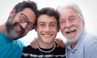 Portrait of handsome people, multigenerational family standing outdoors by the sea, hugging each other and smiling. Grandfather, son and teenage grandson.